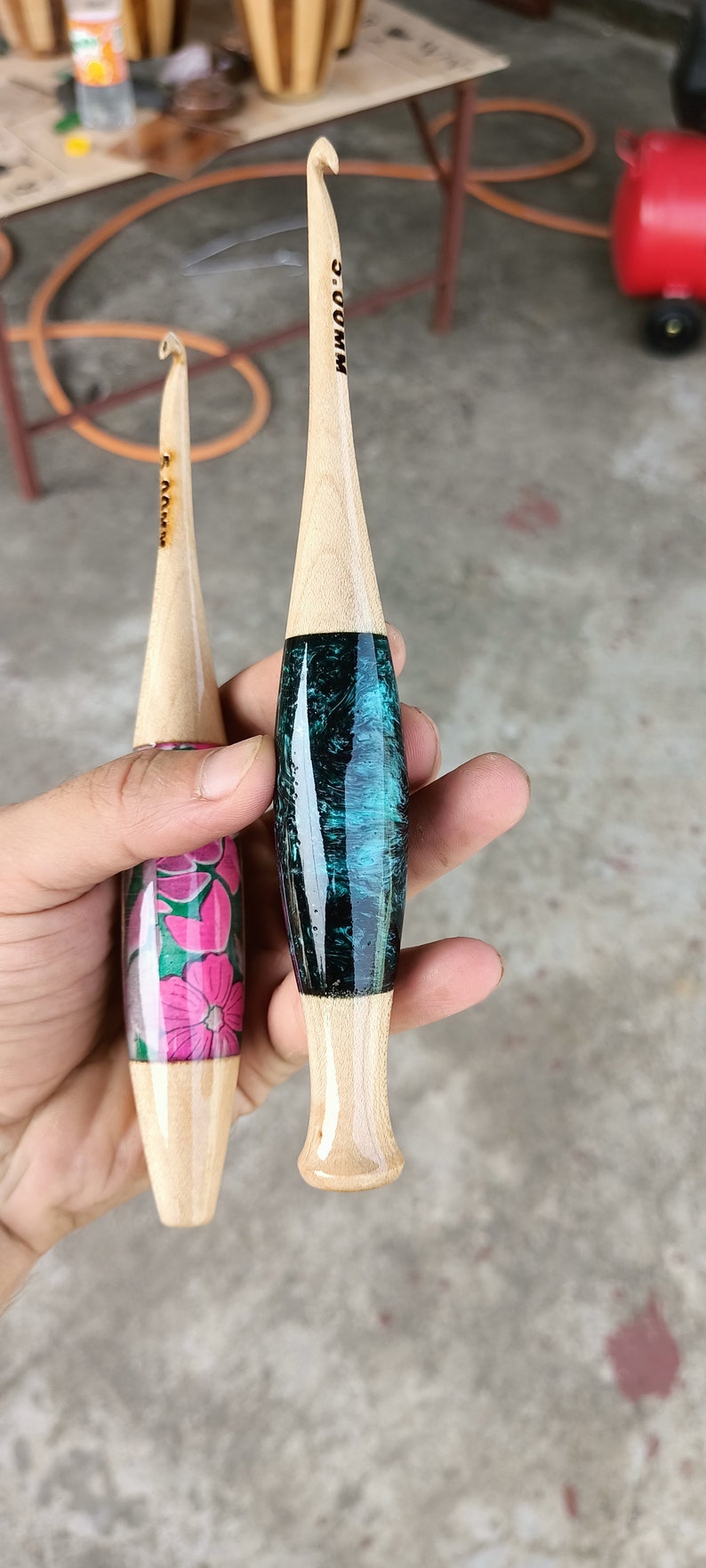 May include: Two wooden crochet hooks with different colored handles. One hook has a teal and black handle, the other has a pink and green floral design. Both hooks have the size marked on them.