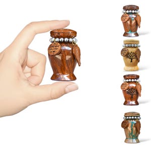 Puede incluir: Una pequeña urna de madera con una hoja decorativa y un acento de cuentas plateadas. La urna tiene una tapa y una pequeña etiqueta de madera que dice "I still need you close to me". Se muestran varias otras urnas.