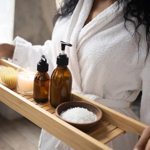 May include: A wooden bath tray holds spa essentials: two amber glass bottles with black pumps, a small wooden bowl of white bath salts, a natural bristle brush, and a lit candle. The tray is being held by a person wearing a white robe.