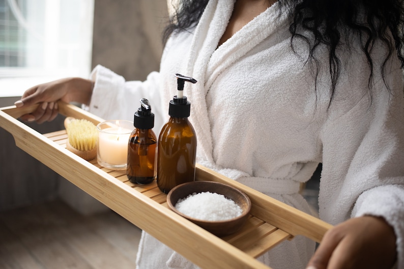 May include: A wooden bath tray holds spa essentials: two amber glass bottles with black pumps, a small wooden bowl of white bath salts, a natural bristle brush, and a lit candle. The tray is being held by a person wearing a white robe.