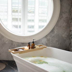May include: A white, freestanding bathtub filled with soapy water and flower petals. A bamboo tray holds two brown bottles, a candle, and a small bowl. A gold faucet and a round window are in the background.