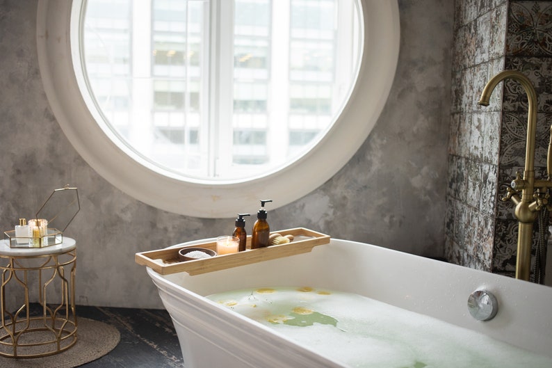May include: A white, freestanding bathtub filled with soapy water and flower petals. A bamboo tray holds two brown bottles, a candle, and a small bowl. A gold faucet and a round window are in the background.