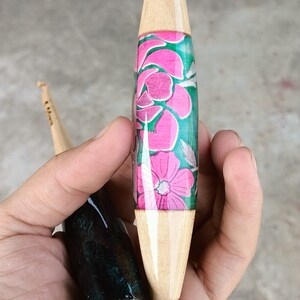 May include: Two wooden crochet hooks with different colored resin handles. One hook has a pink and green floral design, the other has a black and green marbled design.