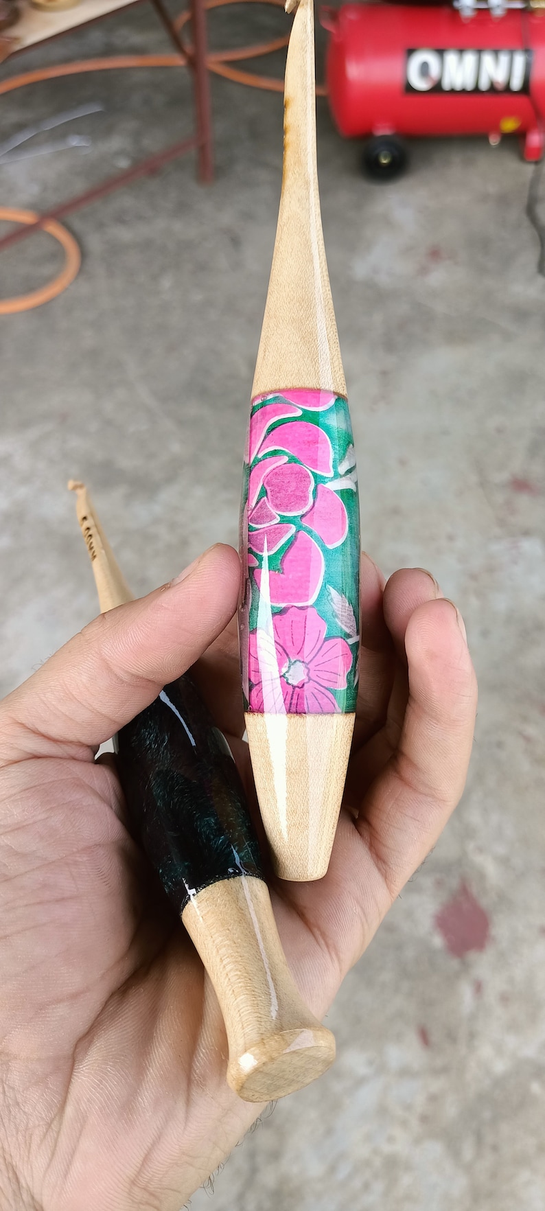 May include: Two wooden crochet hooks with different colored resin handles. One hook has a pink and green floral design, the other has a black and green marbled design.