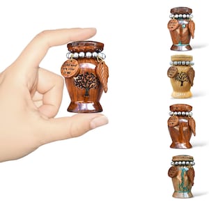 May include: A small, wooden urn with a tree design and a leaf charm. The urn has a dark brown finish, a silver bead accent, and a small wooden tag that reads "I still need you close to me."