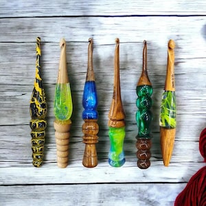 K&ouml;nnte beinhalten: Sieben bunte H&auml;kelnadeln aus Holz mit unterschiedlich farbigem Harzgriffen. Die Nadeln sind in einer Reihe auf einer wei&szlig;en Holzoberfl&auml;che angeordnet.