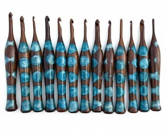 Könnte beinhalten: Eine Reihe von zehn Häkelnadeln aus Holz mit dunkelbraunem Finish und türkisfarbenen Harzakzenten. Die Häkelnadeln sind auf weißem Hintergrund angeordnet, was ihr einzigartiges Design und ihre Handwerkskunst hervorhebt. Jede Nadel hat eine besondere Form.