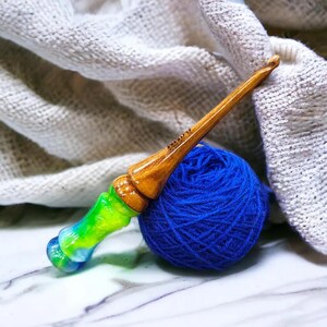 K&ouml;nnte beinhalten: Ein H&auml;kelnadel aus Holz mit einem gr&uuml;nen, blauen und wei&szlig;en Wirbelmuster. Die Nadel liegt auf einer blauen Wollkn&auml;uel und einem wei&szlig;en geh&auml;kelten Stoff.