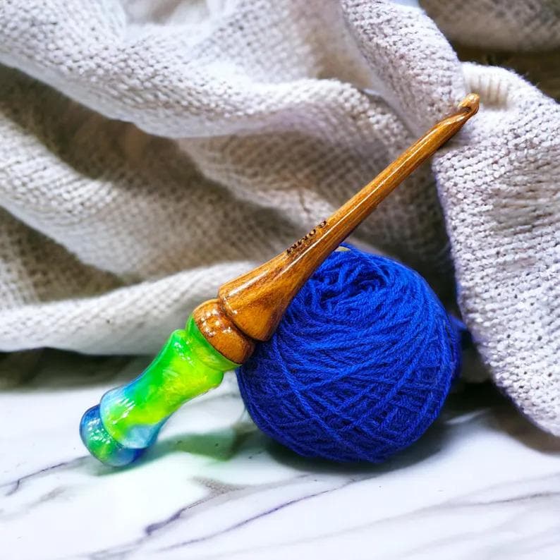 K&ouml;nnte beinhalten: Ein H&auml;kelnadel aus Holz mit einem gr&uuml;nen, blauen und wei&szlig;en Wirbelmuster. Die Nadel liegt auf einer blauen Wollkn&auml;uel und einem wei&szlig;en geh&auml;kelten Stoff.