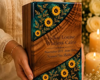 May include: A rectangular wooden memorial box featuring a diagonal inlay of dark teal resin embedded with yellow sunflowers and delicate foliage. The wood surface is engraved with the text Jackie Louise Wheless-Cairo, November 1973 - June 2026, Forever Our Bubbie.