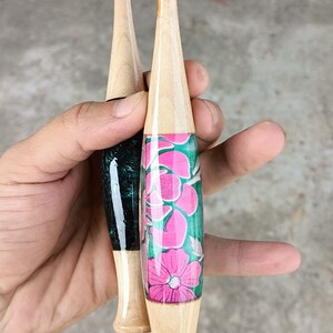 May include: Two wooden crochet hooks with different colored handles. One hook has a black and green handle with the word "SUMMUM" written on it. The other hook has a pink and green floral design on the handle.