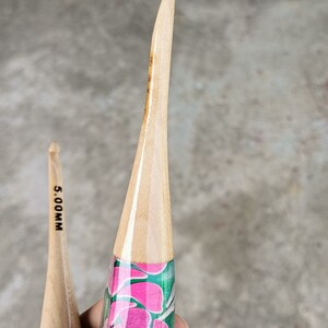 May include: Two wooden crochet hooks with floral designs. The hook on the left is black and green with the text "5.00MM". The hook on the right is green with pink flowers.