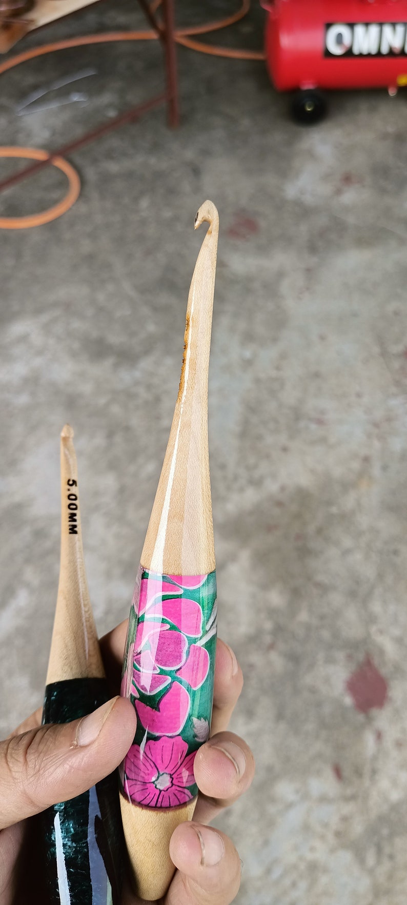 May include: Two wooden crochet hooks with floral designs. The hook on the left is black and green with the text "5.00MM". The hook on the right is green with pink flowers.