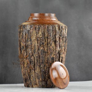 Op de afbeelding: Een houten urn met een schorsachtige textuur en een bijpassend deksel. De urn is handgemaakt en heeft een natuurlijke houtkleur.