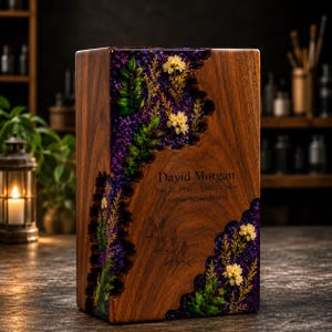May include: A rectangular wooden memorial urn featuring a dark walnut finish with an irregular, jagged inlay of purple resin embedded with dried yellow flowers and green leaves. The front is engraved with David Morgan, July 12, 1950 – April 25, 2024, Forever in our hearts.