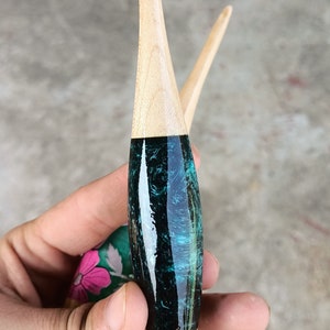 May include: Two wooden crochet hooks with a teal and black marbled design. The hooks are held in a hand.