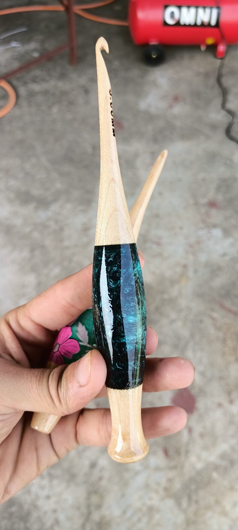 May include: Two wooden crochet hooks with a teal and black marbled design. The hooks are held in a hand.