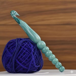 May include: A teal and white marbled crochet hook rests on a ball of dark blue yarn. The hook has a beaded handle and a curved end for catching yarn. The yarn is tightly wound, and the hook is angled across the top of the ball.