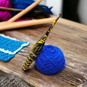 K&ouml;nnte beinhalten: Eine Nahaufnahme eines gelben und schwarzen H&auml;kelhakens mit einem blauen Wollkn&auml;uel. Der Haken ist aus Holz und hat eine strukturierte Oberfl&auml;che. Die Wolle ist weich und flauschig.