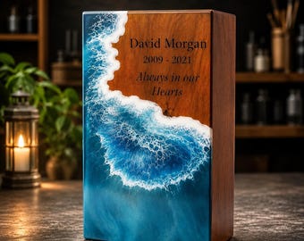 May include: A rectangular wooden cremation urn featuring a resin ocean wave design on the front. The wood is engraved with the text David Morgan, 2009 - 2021, and Always in our Hearts. The resin depicts blue water with white sea foam.