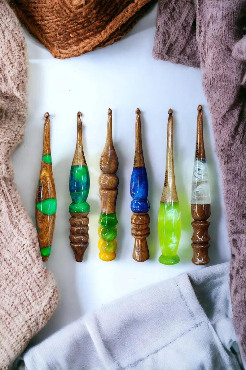 May include: Six wooden crochet hooks with colorful resin handles. The hooks are arranged in a row on a white surface. The colors of the resin handles are green, blue, yellow, and white.