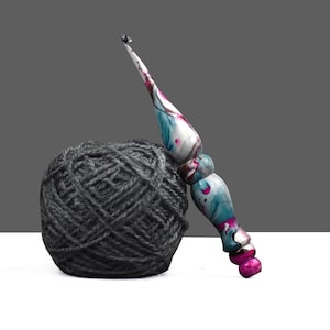 Puede incluir: Una madeja de lana gris con un ganchillo colorido. El ganchillo tiene un diseño veteado con tonos de azul, rosa y blanco. El ganchillo está apoyado en la madeja de lana, que está hecha de hilo gris.