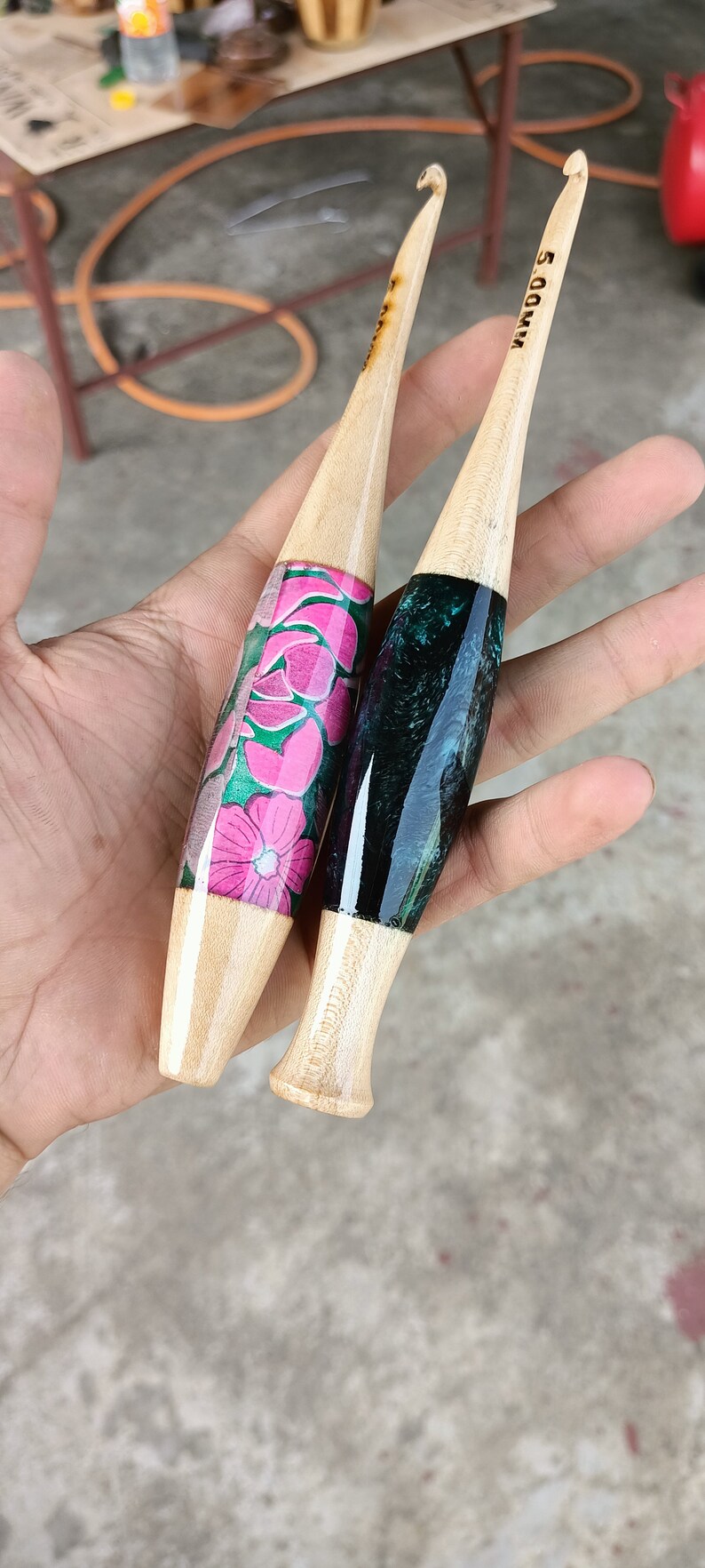 May include: Two wooden crochet hooks with different colored resin handles. One hook has a pink and green floral design, the other has a black and green marbled design. The hooks are labeled "5.00 MM" and "5.00 MM".