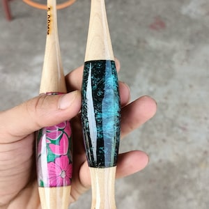 May include: Two wooden crochet hooks with different colored handles. One hook has a teal and black handle, the other has a pink and green floral design. Both hooks have the size marked on them.
