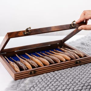 Puede incluir: Una caja de madera con tapa de cristal, que muestra una colección de ganchillos de madera. Los ganchillos están dispuestos sobre un forro de terciopelo azul. La caja está abierta, revelando el contenido. La madera tiene un acabado cálido y natural.