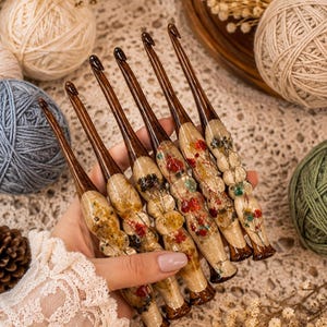 May include: A set of five crochet hooks with dark brown tapered tips and ergonomic handles made of clear resin. Each handle is embedded with various dried flowers in shades of red, yellow, green, and white, creating a unique floral pattern.