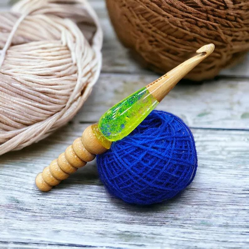 K&ouml;nnte beinhalten: Ein H&auml;kelnadel aus Holz mit einem gr&uuml;nen und gelben Harzgriff. Die Nadel liegt auf einer blauen Garnrolle.