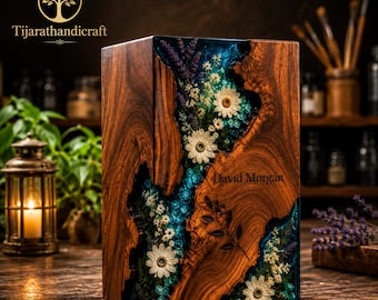 May include: A rectangular wooden urn featuring a central river-like inlay of translucent blue resin embedded with dried white daisies, lavender sprigs, and green foliage. The name David Morgan is engraved on the wood surface.