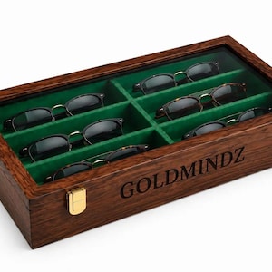 Può includere: Una custodia rettangolare in legno con coperchio in vetro e interno in velluto verde, dotata di sei scomparti per occhiali. La parte anteriore della scatola in legno marrone scuro presenta l'incisione GOLDMINDZ e due chiusure metalliche dorate.