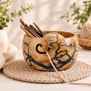 May include: A round wooden yarn bowl featuring a natural grain finish with inlaid blue resin patterns. The bowl has a spiral cutout for yarn and contains a ball of beige yarn along with several wooden crochet hooks resting inside the container.