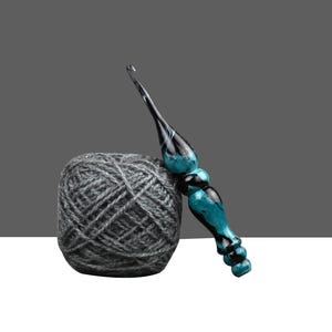 May include: A ball of gray yarn next to a crochet hook. The hook has a teal and black handle. The yarn is a tightly wound ball of gray thread. The background is a gradient of gray and white.