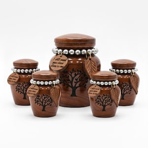 May include: A set of wooden urns in different sizes, featuring a dark brown finish and a tree design. The urns are decorated with silver bead accents and leaf-shaped charms. Each urn has a wooden tag with the text "I will need you close to me."