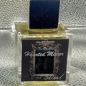 May include: A clear glass perfume bottle with a black square cap. The label reads "JULIET ROSE Haunted Mirror 50ml" in white script. The bottle contains a golden liquid.