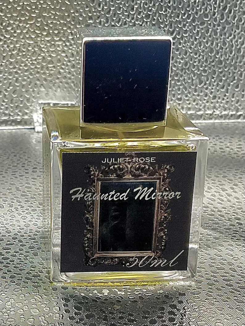 May include: A clear glass perfume bottle with a black square cap. The label reads "JULIET ROSE Haunted Mirror 50ml" in white script. The bottle contains a golden liquid.
