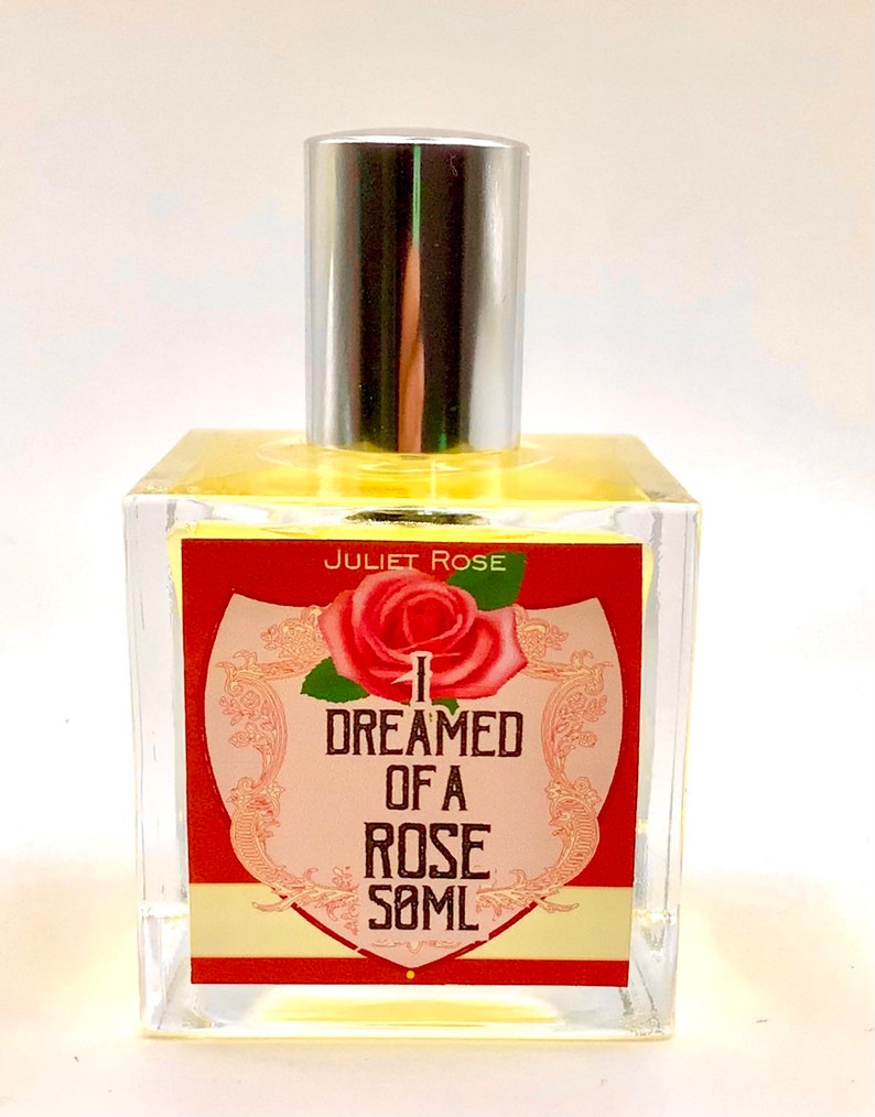 Puede incluir: Una botella de vidrio transparente con una tapa plateada y una etiqueta roja. La etiqueta presenta una rosa rosa y el texto "I DREAMED OF A ROSE 50ML" en letras negras. El texto "JULIET ROSE" est&aacute; impreso en negro en la parte superior de la etiqueta.