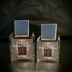 May include: Two square glass perfume bottles with dark blue caps. The labels read "JULIET ROSE Haunted Mirror 50ml" with a framed image. The bottles are on a dark surface.