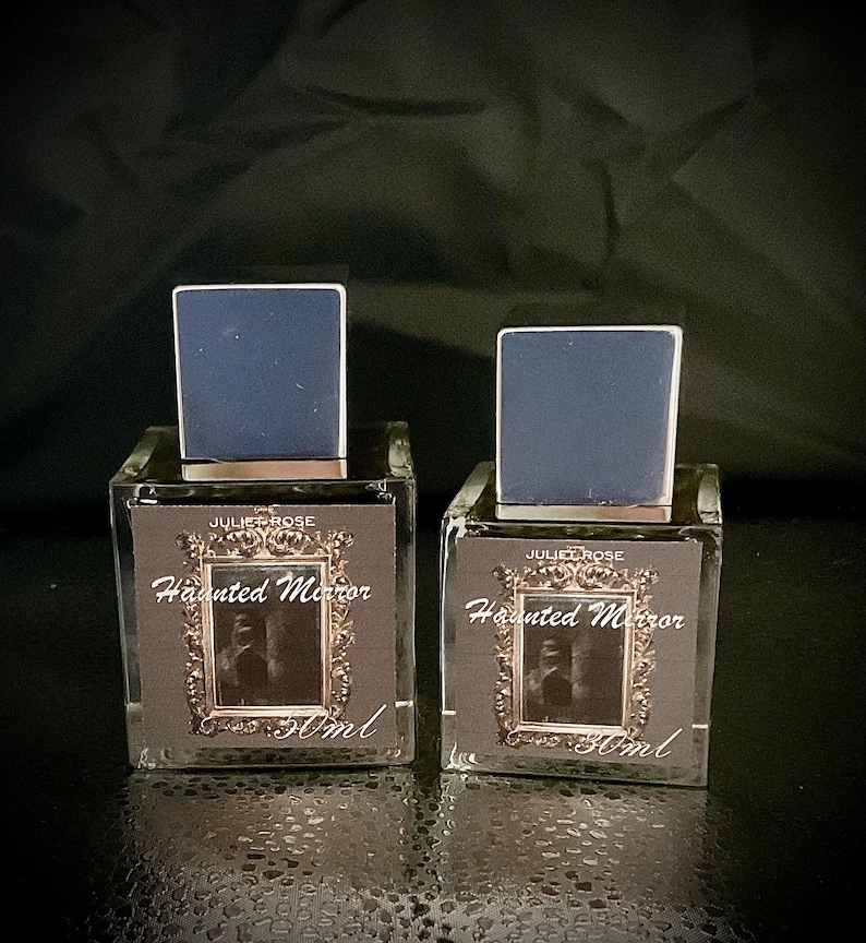May include: Two square glass perfume bottles with dark blue caps. The labels read "JULIET ROSE Haunted Mirror 50ml" with a framed image. The bottles are on a dark surface.