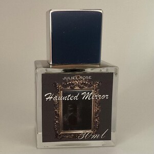 May include: A clear glass perfume bottle with a dark blue cap and silver trim. The label reads "JULIET ROSE Haunted Mirror" with a decorative frame and the text "30ml". The bottle is square and sits on a white surface.