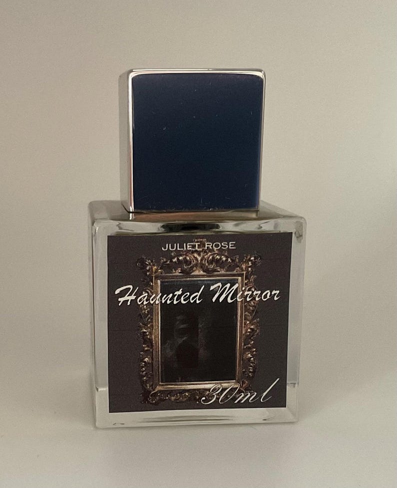 May include: A clear glass perfume bottle with a dark blue cap and silver trim. The label reads "JULIET ROSE Haunted Mirror" with a decorative frame and the text "30ml". The bottle is square and sits on a white surface.