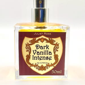 May include: A clear glass perfume bottle with a silver cap. The label reads "Juliet Rose Dark Vanilla Intense Eau de Parfum 50ml" in gold lettering on a burgundy background. The perfume inside is a golden colour.