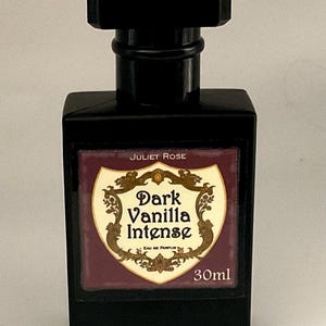 May include: A black rectangular perfume bottle with a black cap. The label reads "Juliet Rose Dark Vanilla Intense" with a decorative design. The bottle contains 30ml of perfume.