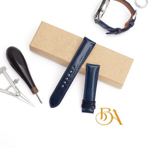 May include: Two blue leather watch straps with white stitching. The straps are on a brown cardboard box. A watch with a blue leather strap is in the background.