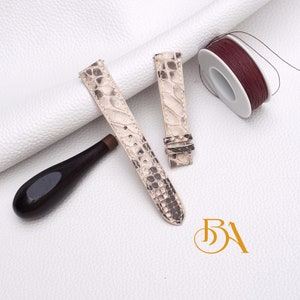 Op de afbeelding: Twee beige leren horlogebandjes met een slangenhuidpatroon. De bandjes liggen op een wit oppervlak met een spoel rood garen en een houten gereedschap.