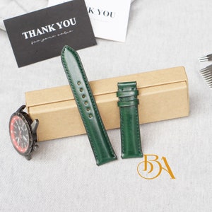 May include: Two green leather watch bands in a brown box. The bands are different sizes, one is longer than the other. The bands are both a dark green color.
