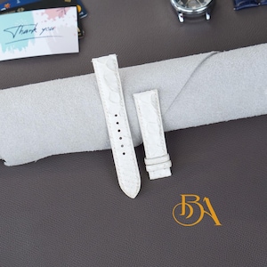 Pode incluir: Pulseira de relógio branca com um padrão texturizado. A pulseira tem uma série de orifícios para ajuste e uma fivela. A pulseira do relógio é mostrada em uma superfície cinza com um pano branco dobrado.