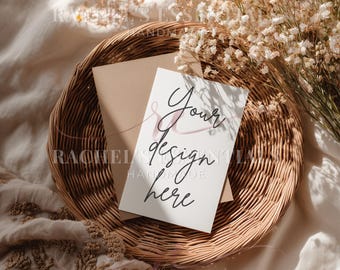 May include: A white card with the text "Your design here" rests on a brown envelope inside a woven basket. Dried white flowers and a beige fabric surround the basket. The image has a soft, natural aesthetic.